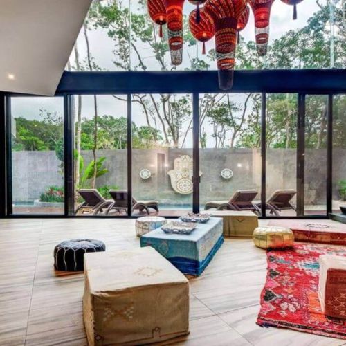 Area of the house decorated with cushions, carpets, and lounge chairs with a view through the open glass doors to the pool
